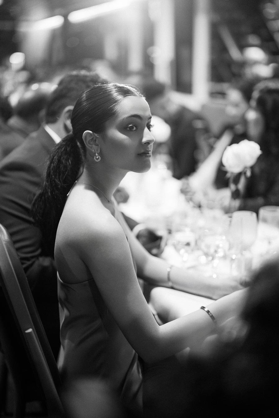 Black and white photo of a woman in formal attire at a banquet, sitting at a table set with glasses and flowers, with soft lighting highlighting her features, capturing an elegant event atmosphere