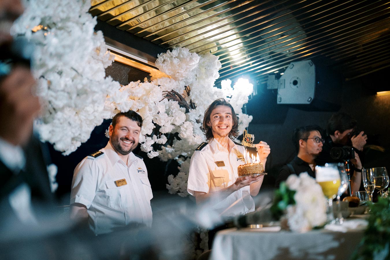 Crew members celebrating with a festive cake onboard, surrounded by floral decorations and smiling for a special occasion.