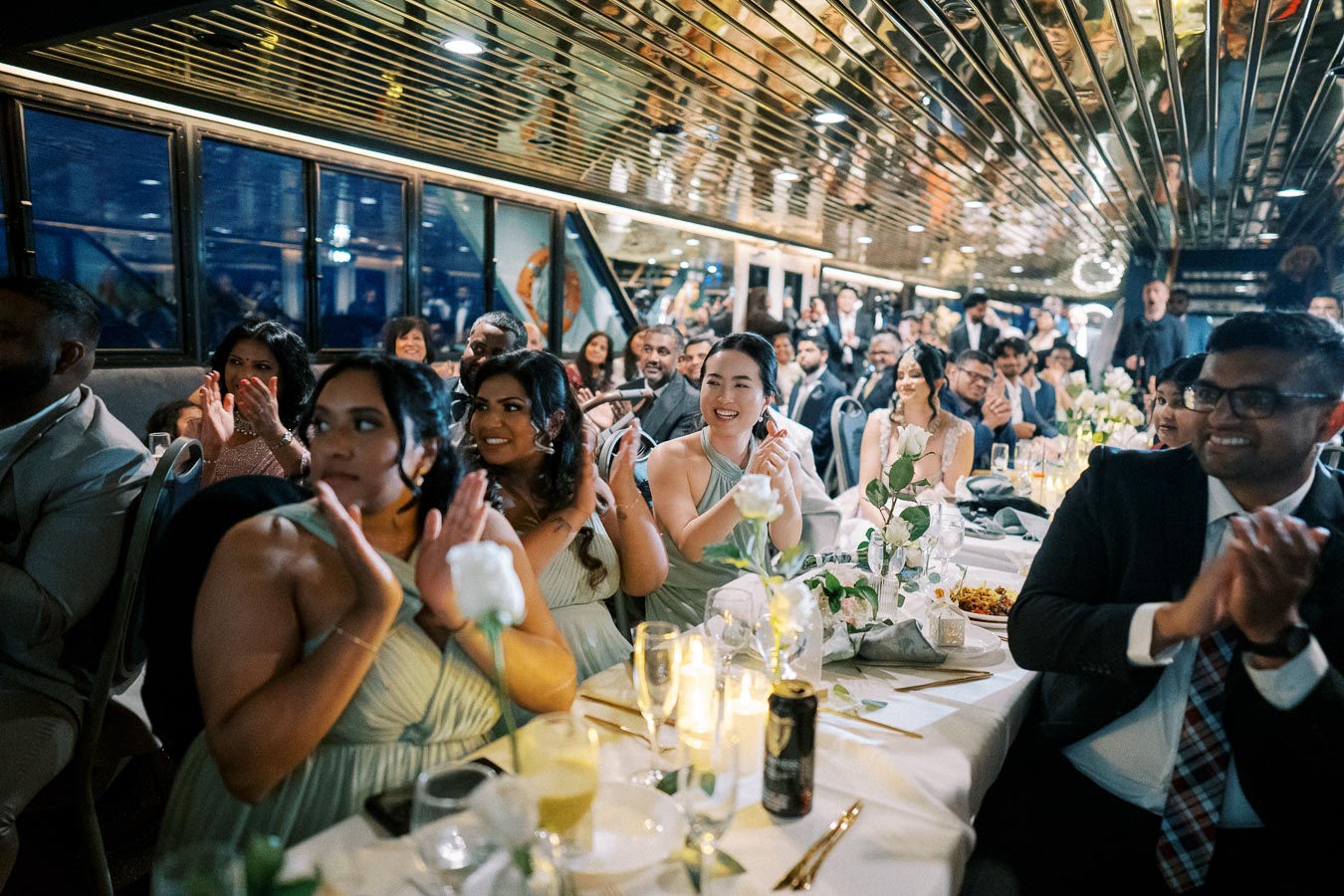 Guests at a wedding reception on a boat, seated around elegantly decorated tables with flowers and candles, smiling and clapping during a celebration.