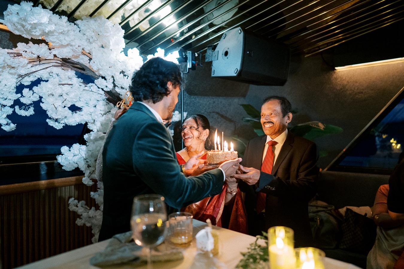 Elder couple celebrating a birthday, receiving a lit birthday cake from a younger man in a warmly lit room with elegant floral decorations.