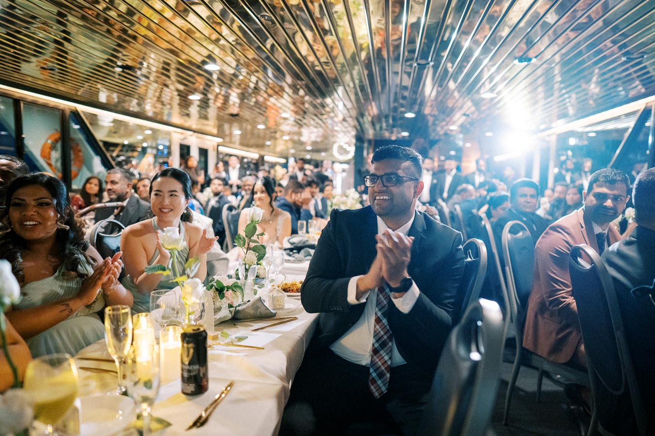 Guests in formal attire clapping and smiling at a wedding reception, sitting at elegantly decorated tables with flowers and candles, under a mirrored ceiling.