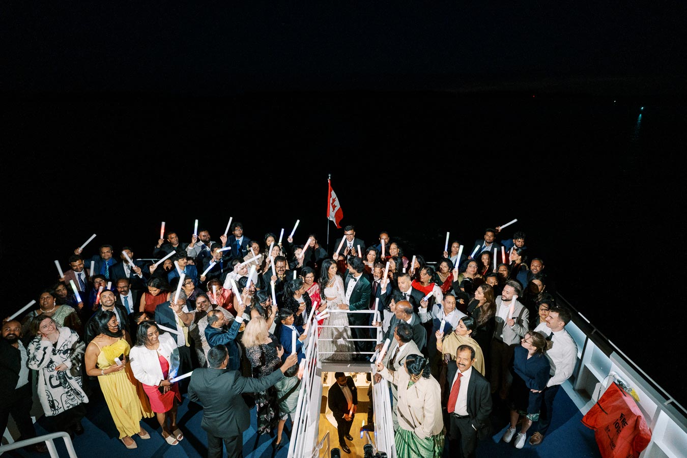 A large group of people, dressed in formal and colorful attire, gather on a boat at night under a dark sky. They are holding illuminated sticks and standing near a Canadian flag, creating a festive and celebratory atmosphere.