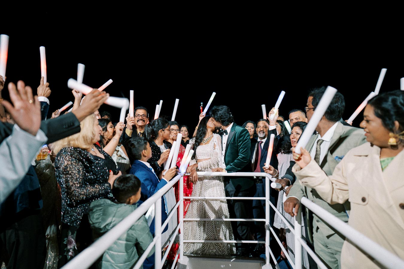 A bride and groom share a kiss surrounded by cheering guests holding light sticks at a nighttime wedding celebration on a boat deck.