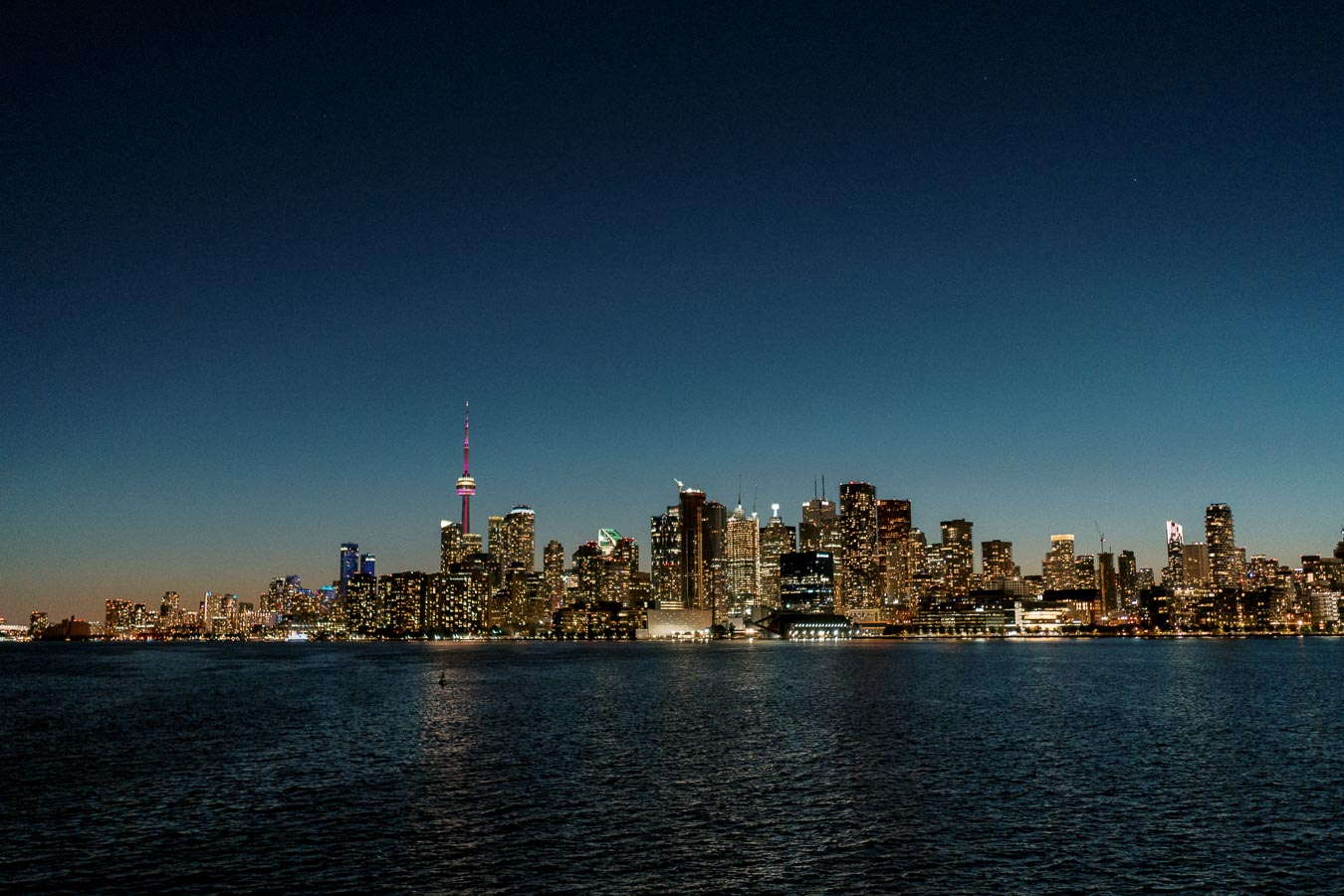 Skyline view of Toronto at dusk with illuminated CN Tower and city lights reflecting on Lake Ontario.