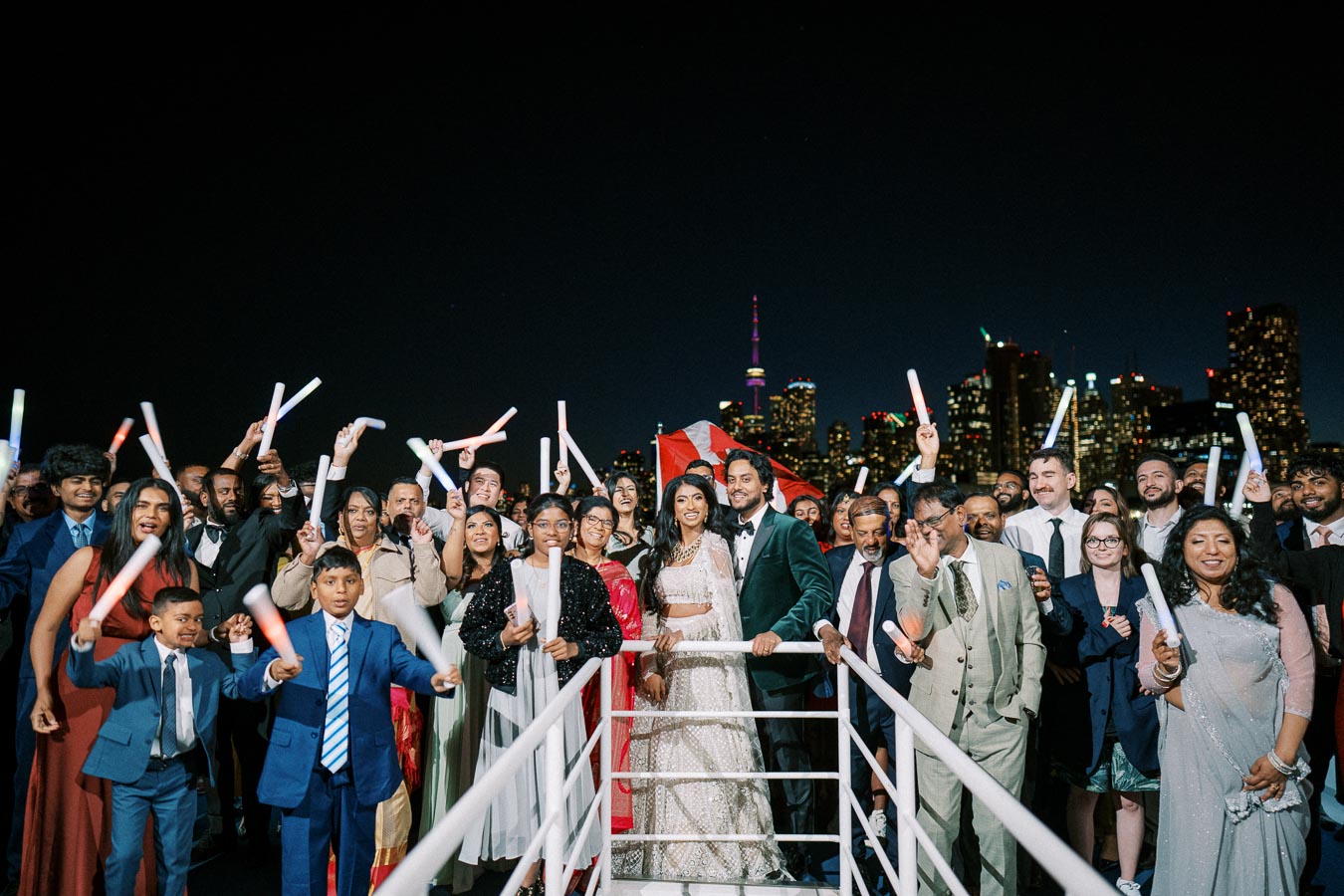 Group of people celebrating at a nighttime event on a boat with glowing sticks, with a city skyline illuminated in the background.