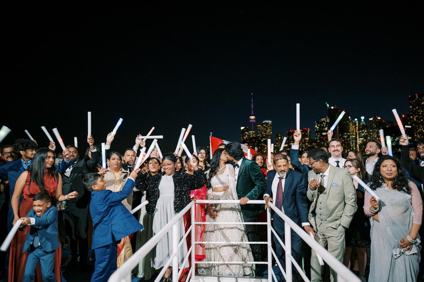 A joyful wedding celebration takes place at night with a diverse group of guests holding glow sticks aboard a boat, against a backdrop of a vibrant city skyline. The bride and groom share a romantic kiss in the center, surrounded by family and friends.
