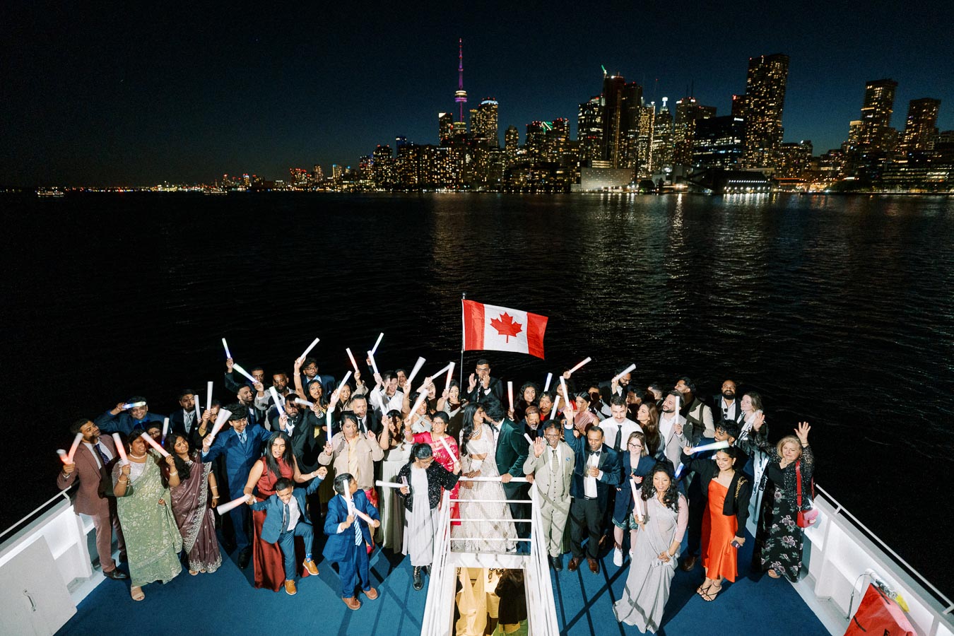 A large group of people celebrating on a boat at night with a Canadian flag, with the Toronto skyline illuminated in the background.