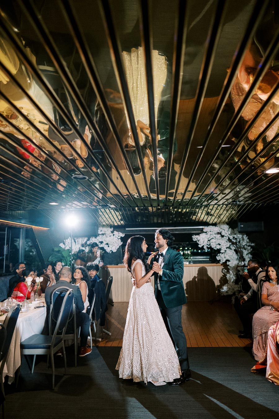 Couple sharing a dance at an elegant wedding reception with a reflective ceiling, surrounded by seated guests and floral decorations.
