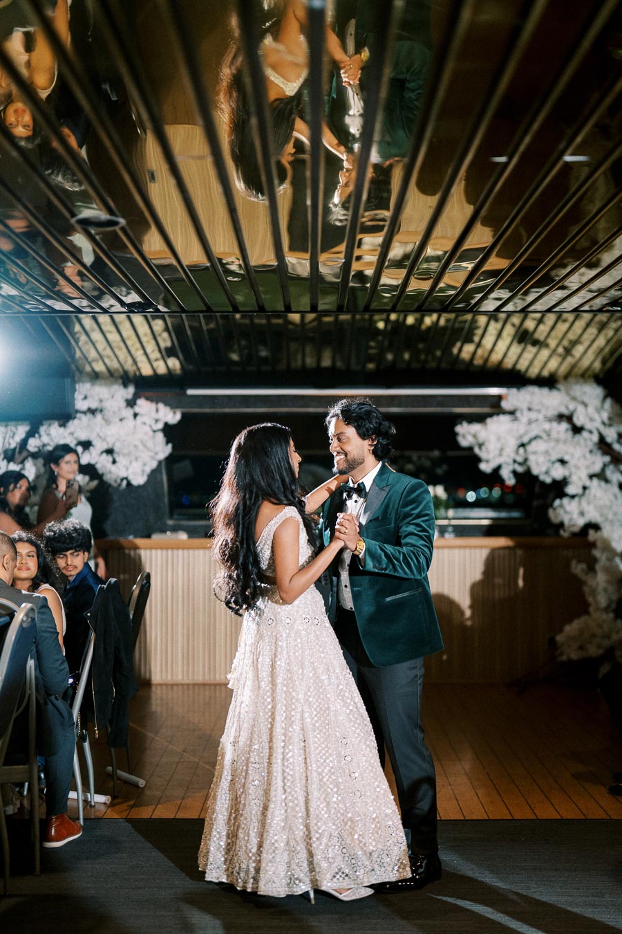 Couple dancing at a wedding reception, with the bride in a sparkling white gown and the groom in a dark suit, reflected on the ceiling, surrounded by guests seated at tables and floral decorations in the background.