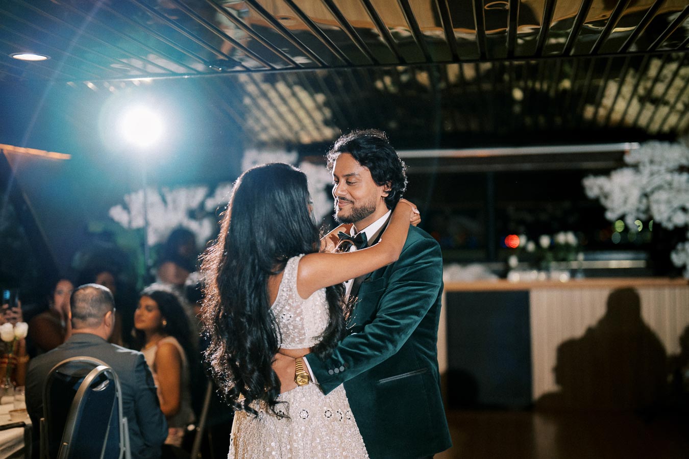 A couple sharing a romantic dance at a wedding reception, with the bride in a sparkling white gown and the groom in a dark velvet suit.