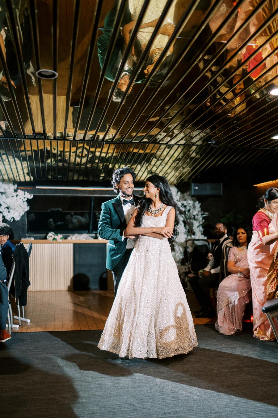 Happy couple dancing at a wedding reception, with a mirrored ceiling reflecting their joyful expressions, amidst elegantly dressed guests and floral decor.
