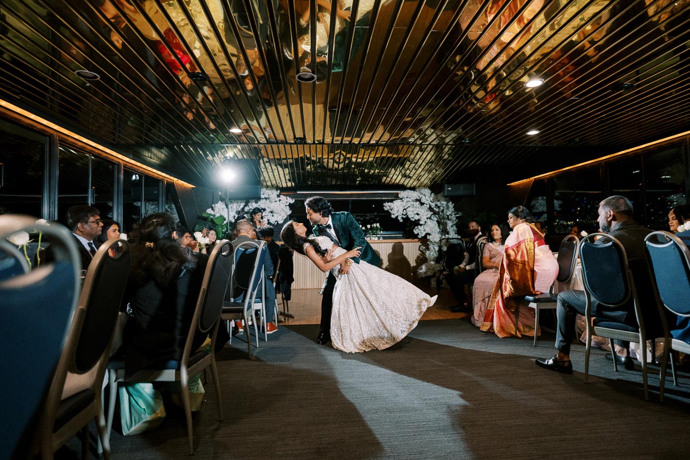 A couple performing a romantic dance in an elegant venue, surrounded by seated guests watching attentively, with decorative floral arrangements in the background.