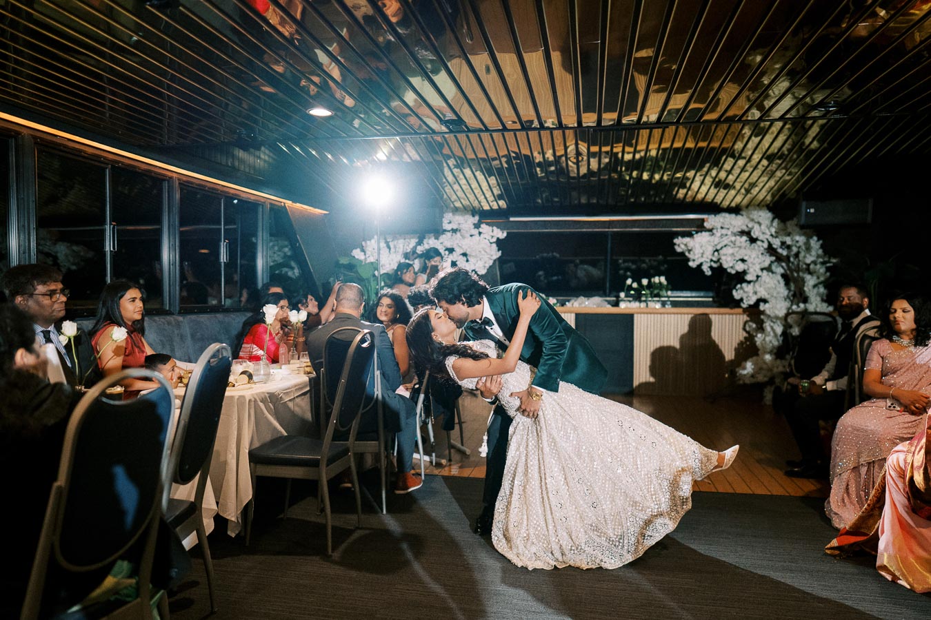 A couple shares a romantic dance, the groom in a green suit and the bride in an elegant white gown, surrounded by guests at a beautifully decorated indoor wedding reception.
