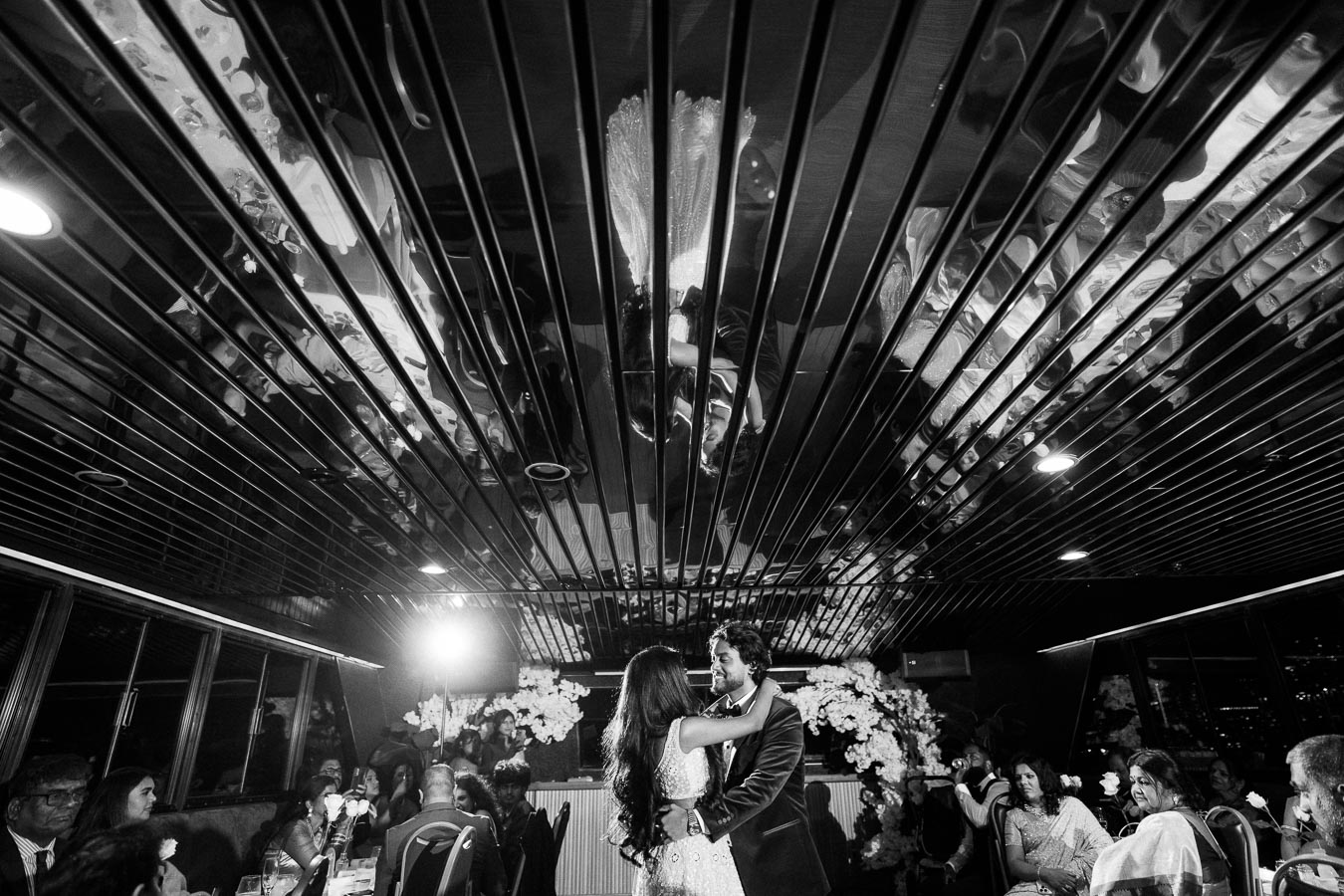 A couple dances closely at a wedding reception, reflected in a mirrored ceiling, surrounded by guests seated at tables decorated with flowers.