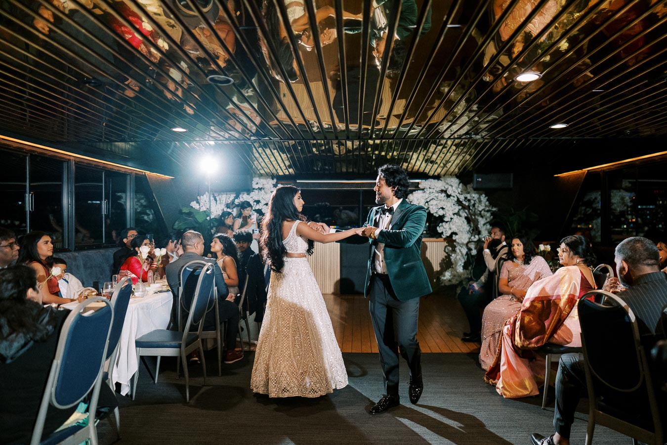 A couple elegantly dances in a warmly lit wedding reception venue, surrounded by seated guests at round tables. The woman wears a sparkling white gown, and the man is in a dapper suit. A floral backdrop and reflections on the ceiling add to the celebratory ambiance.
