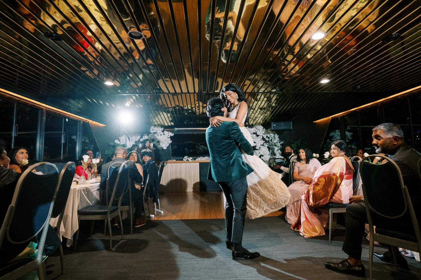 A joyful couple dancing at their wedding reception in an elegantly decorated venue with guests seated around, capturing the ambiance of celebration and love.