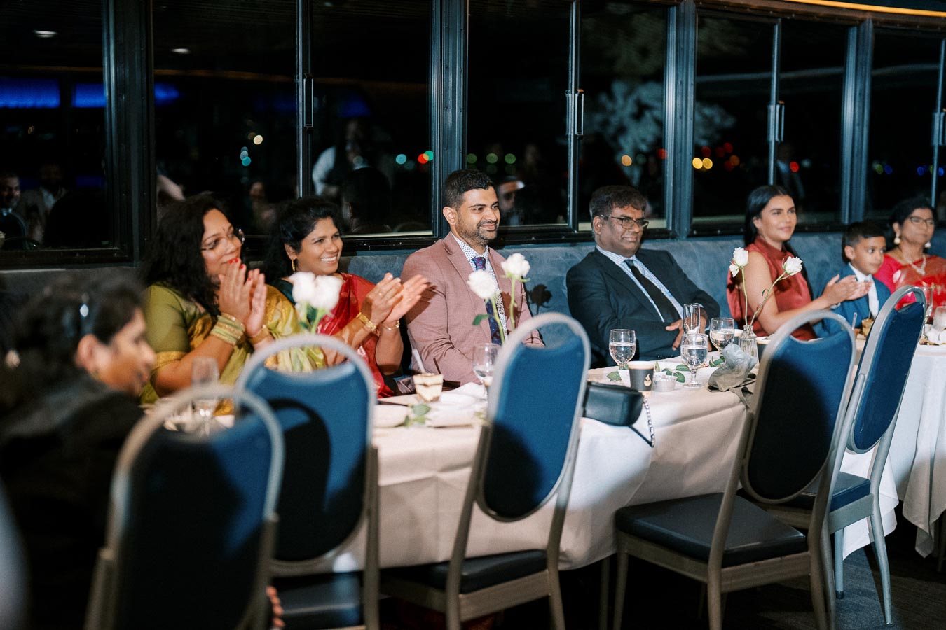 A group of people sitting at a formal dinner event, clapping and smiling, with elegant table settings and floral decorations.