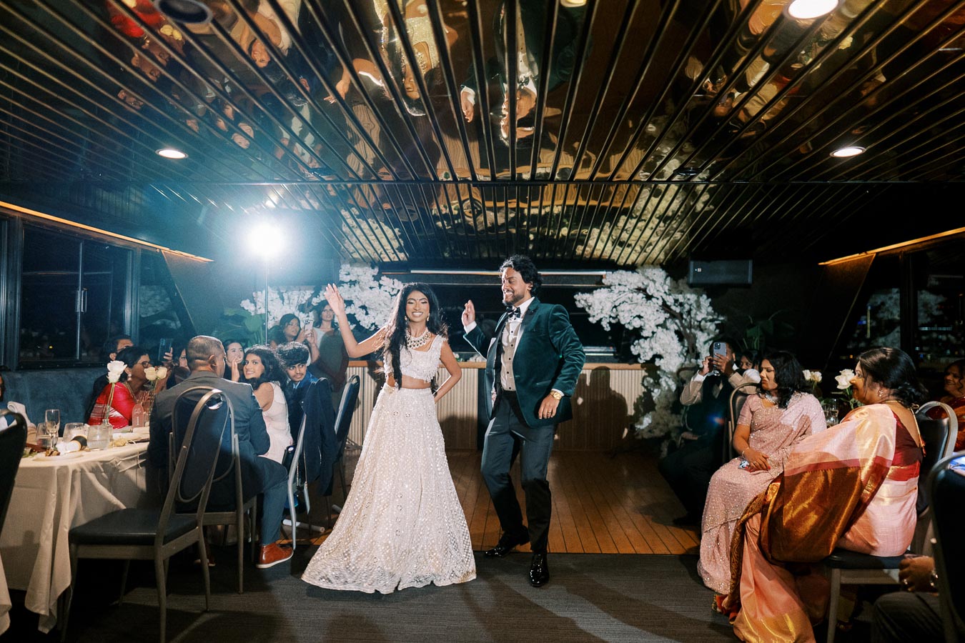 Guests enjoying a lively wedding dance celebration in an elegant venue, featuring a couple dancing joyfully in formal attire under warm lighting with reflections on a sleek ceiling.