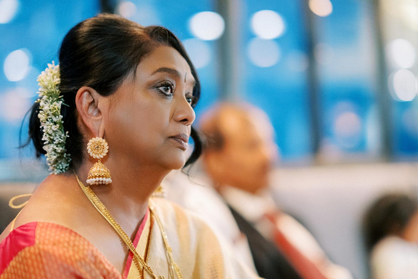 Senior woman wearing traditional Indian attire and jewelry, with hair adorned with flowers, attentively listening at an event in a blurred background setting.
