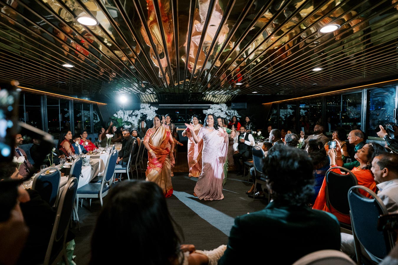 A group of women in elegant sarees perform a dance at a festive indoor event, with an audience seated at tables watching attentively, capturing the celebration on their smartphones.