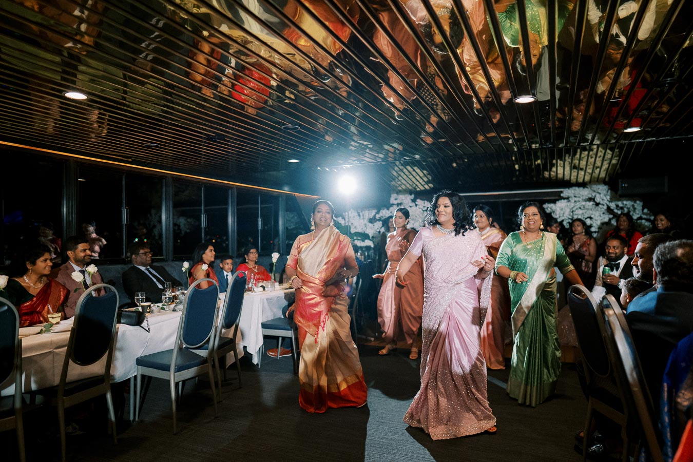 A group of elegantly dressed women and guests at a lively indoor event, featuring colorful saris and traditional attire, with smiling faces and decorative lighting enhancing the festive atmosphere.