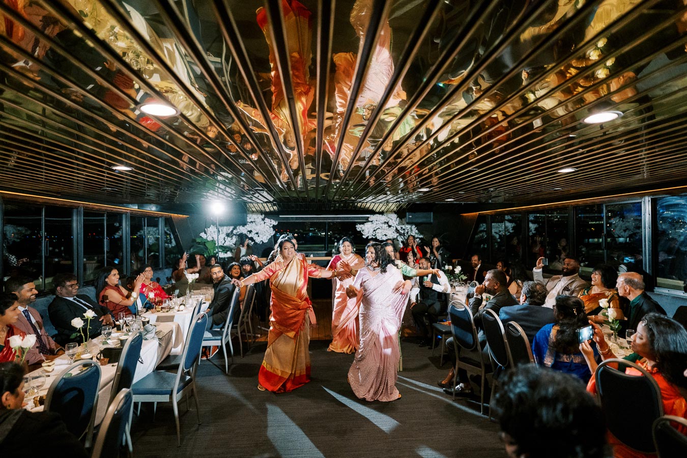 Elegant indoor wedding celebration on a boat, featuring guests in formal attire, beautifully set dining tables, and three women joyfully dancing in vibrant saris, under a reflective ceiling with dim lighting.