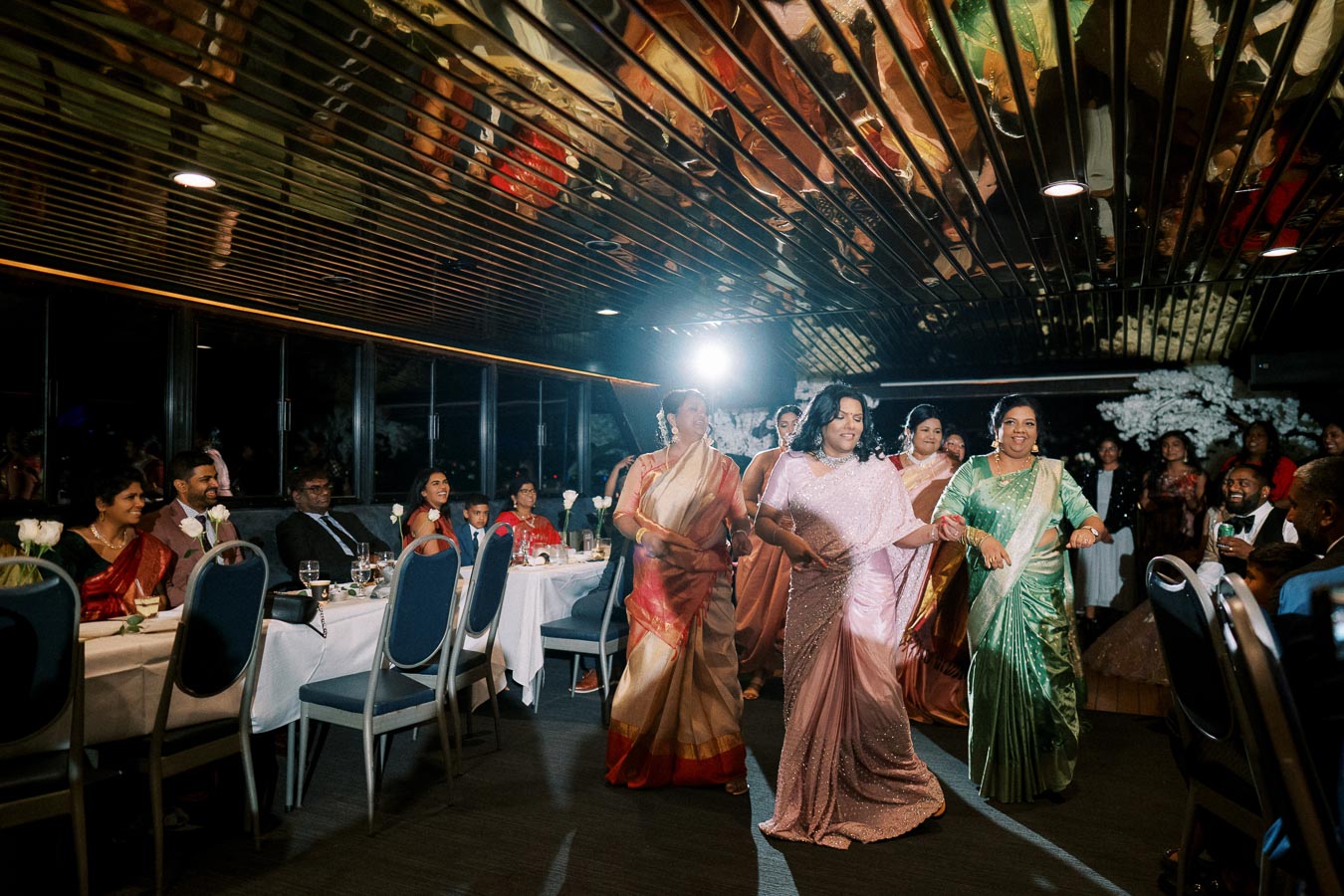 A group of women in colorful traditional sarees dancing joyfully at an indoor celebration, with guests seated at tables watching and a reflective ceiling above.