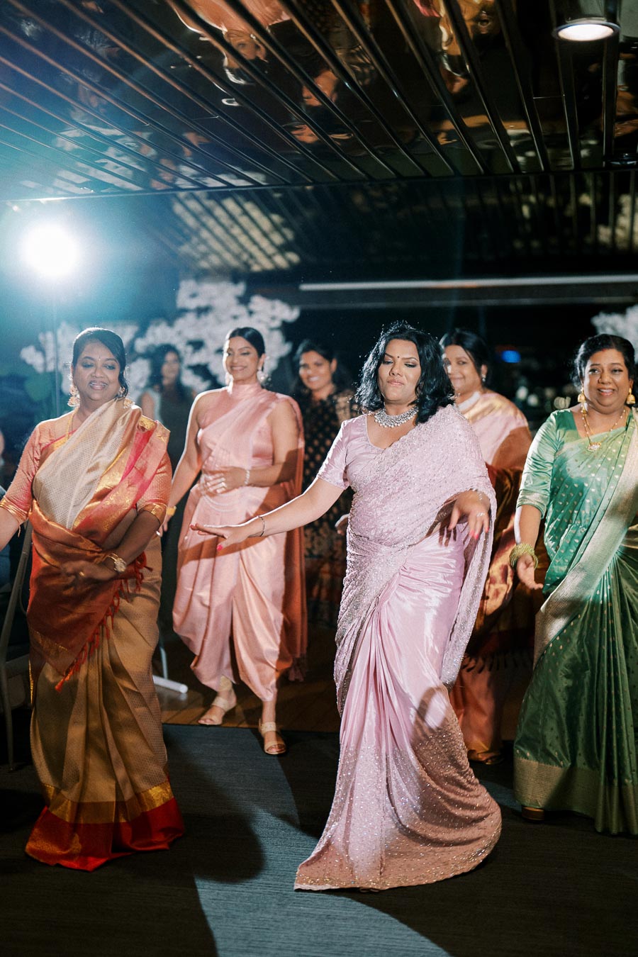 A group of women in elegant sarees dancing joyfully at an indoor event, with smiles and vibrant colors enhancing the festive atmosphere.