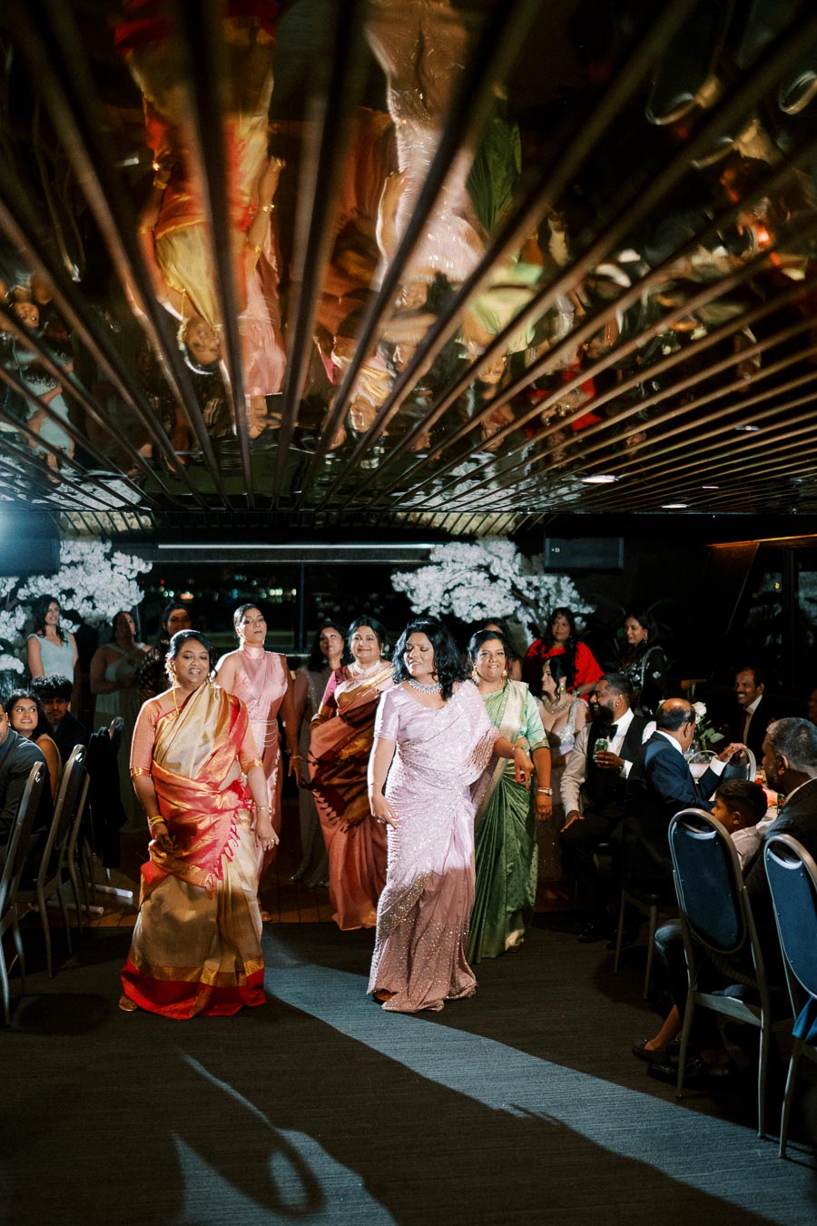 A group of women in colorful sarees dancing at a festive indoor event with a mirrored ceiling and seated guests in the background.