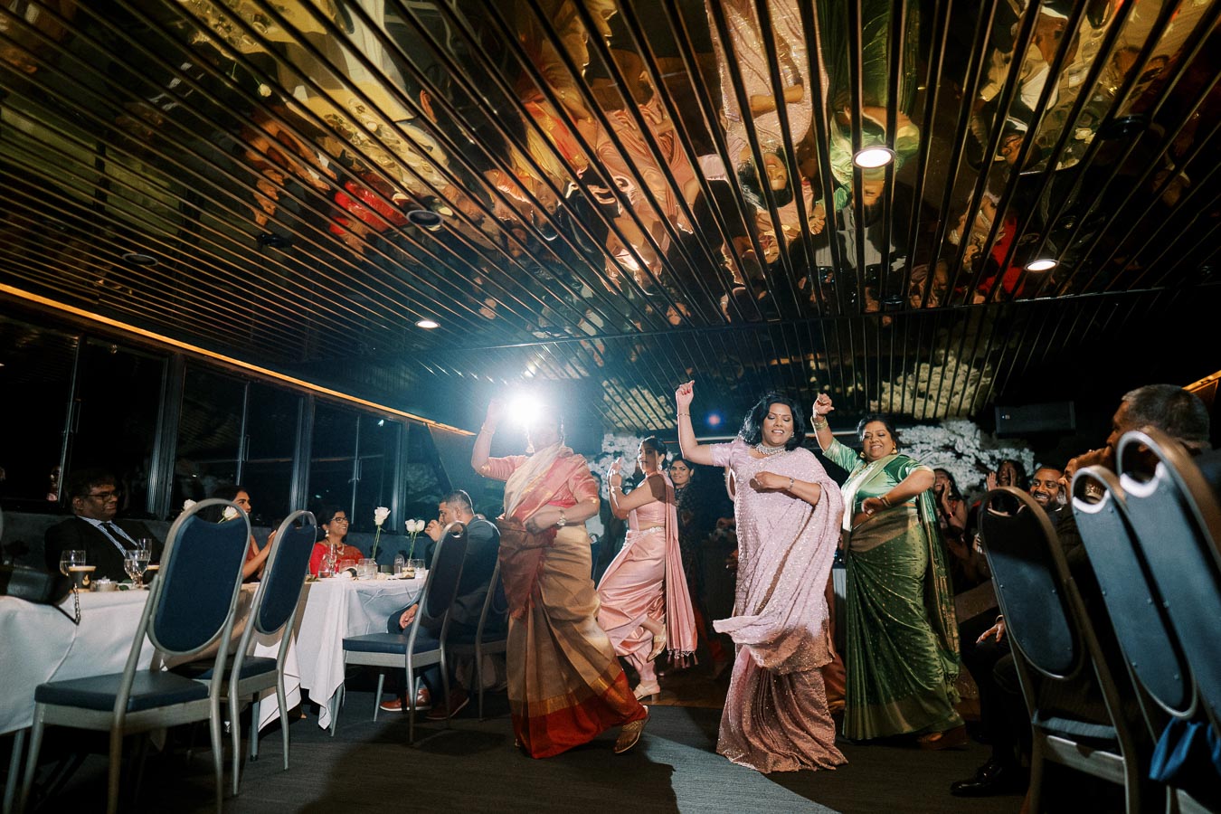 A group of people dressed in traditional Indian attire dancing joyfully at an indoor celebration, with reflections on the ceiling and seated guests watching in the background.