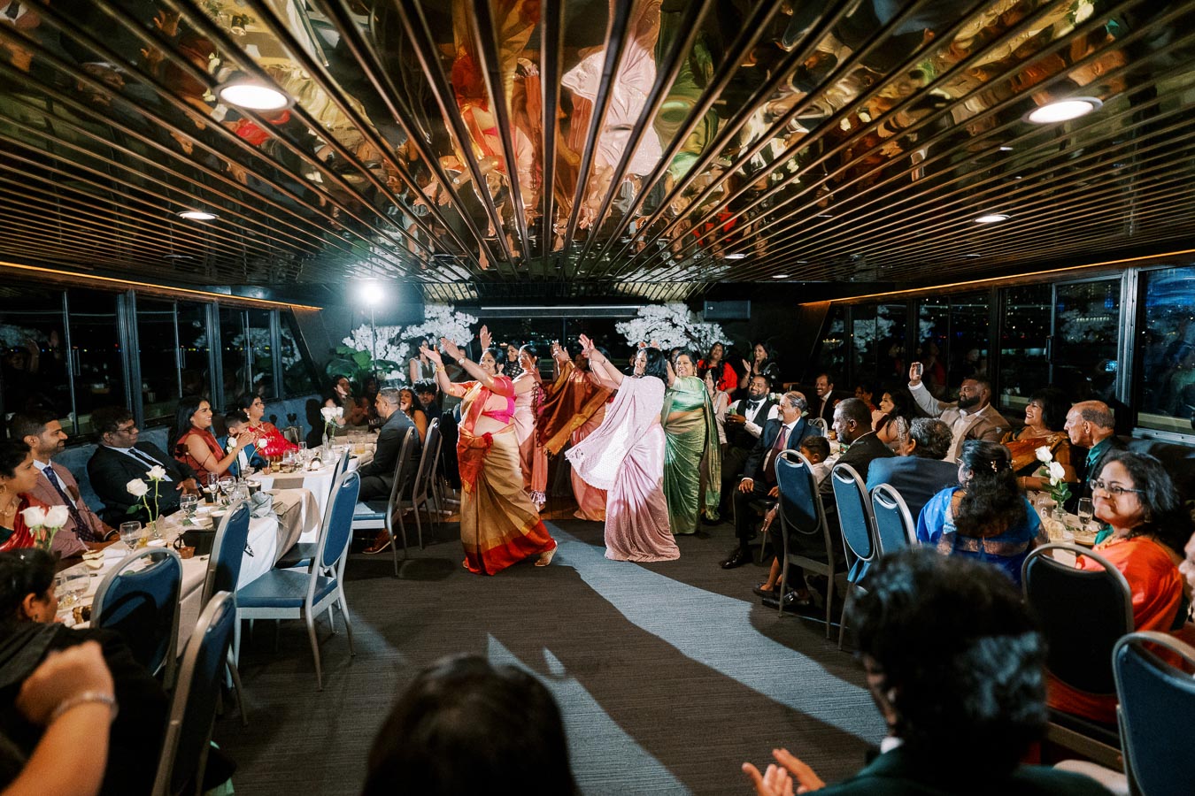 A group of women in colorful traditional attire dance energetically in the center of a festive banquet hall, surrounded by seated guests who watch and applaud their performance.