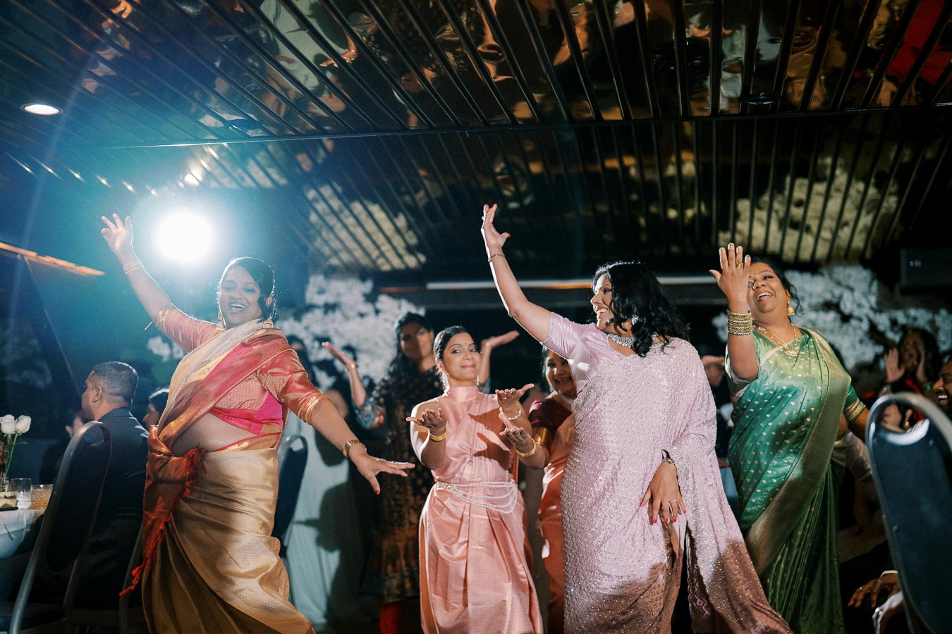A group of women joyfully dancing in traditional colorful saris at a festive indoor event, with dim lighting and a lively atmosphere.