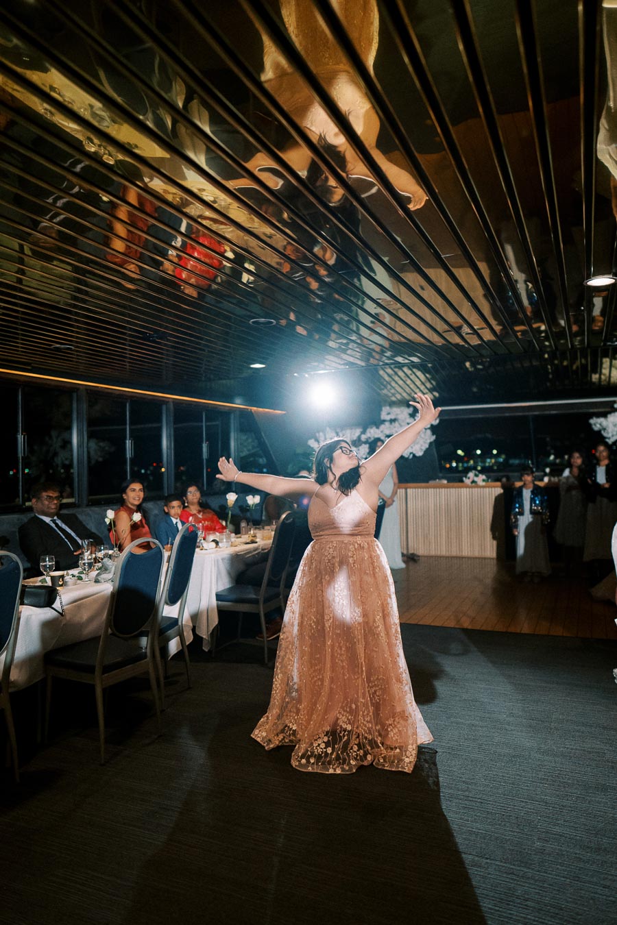 A person in an elegant, flowing gown joyfully dancing at a formal event, with guests seated at tables observing the performance under a reflective ceiling.