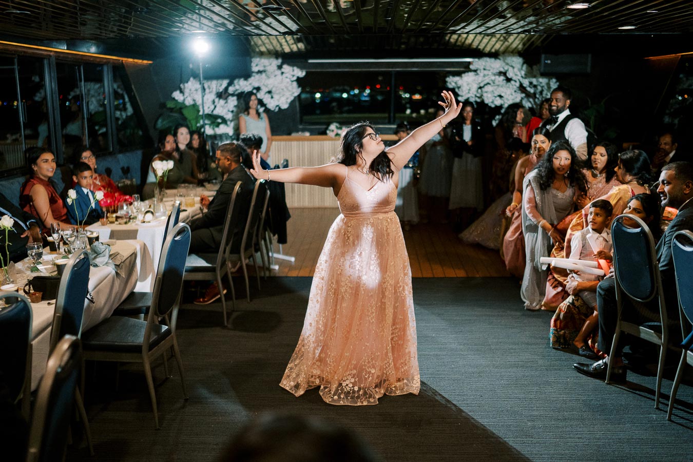 A woman in a pink gown joyfully performs a dance at an event, surrounded by seated guests watching with smiles in a warmly lit venue.