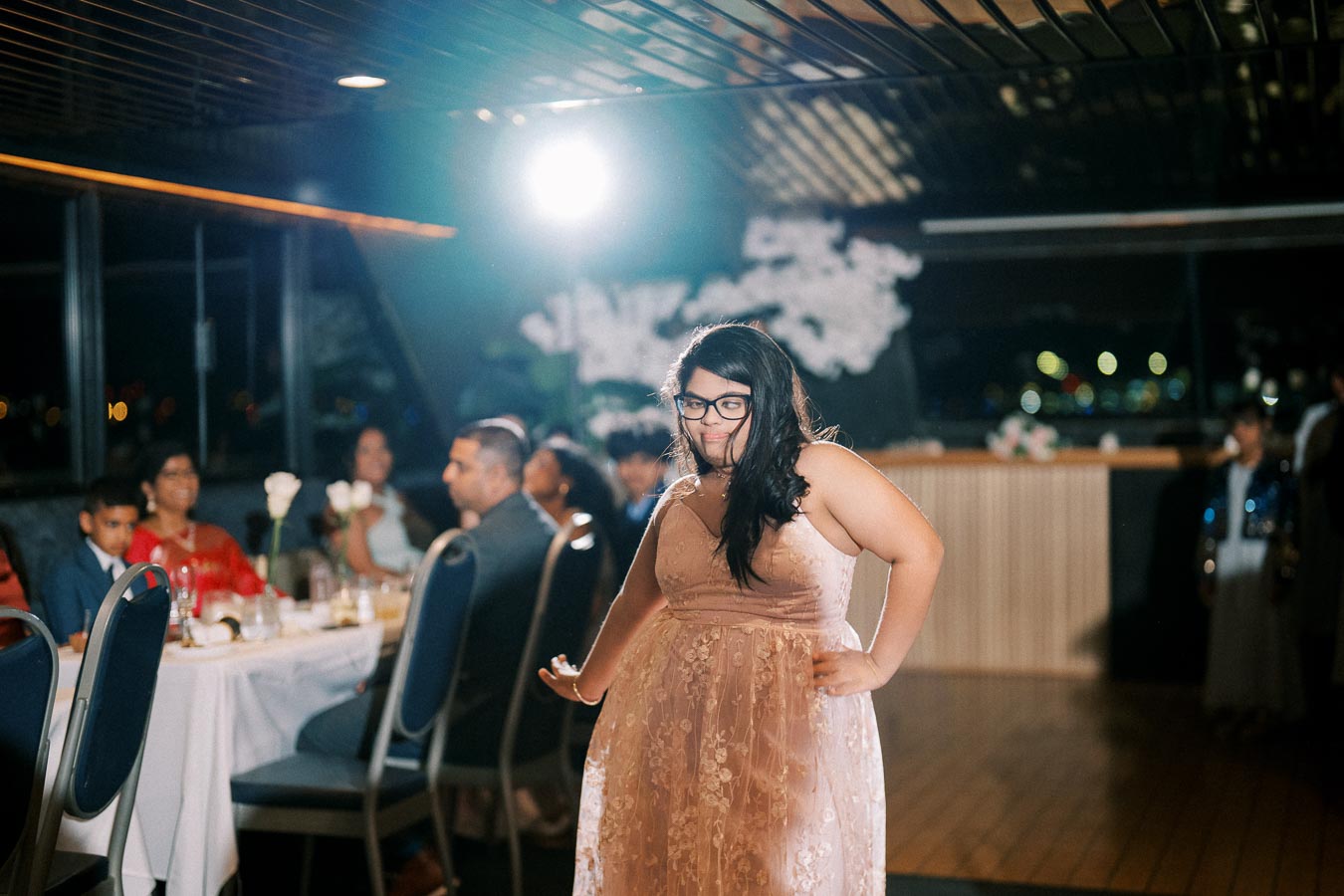 A woman in a stylish pink dress stands confidently in front of a banquet table at a formal event, surrounded by seated guests.