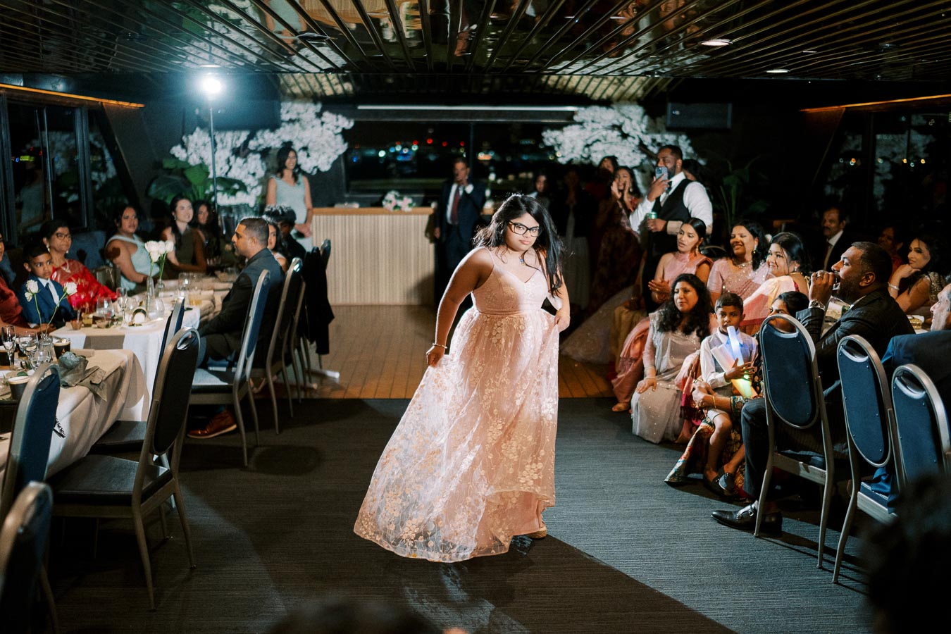 A young woman in a pink gown confidently walks down the aisle at a formal event, surrounded by seated guests. The atmosphere is celebratory with elegant decorations and dim lighting.