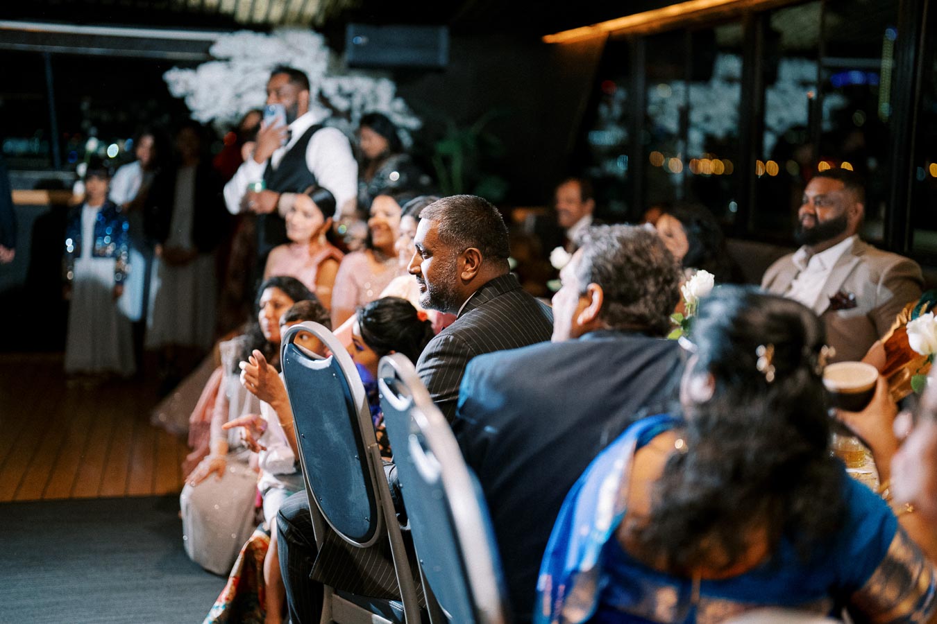 Group of people gathered at a formal event, attentively listening and seated around a table adorned with floral arrangements.