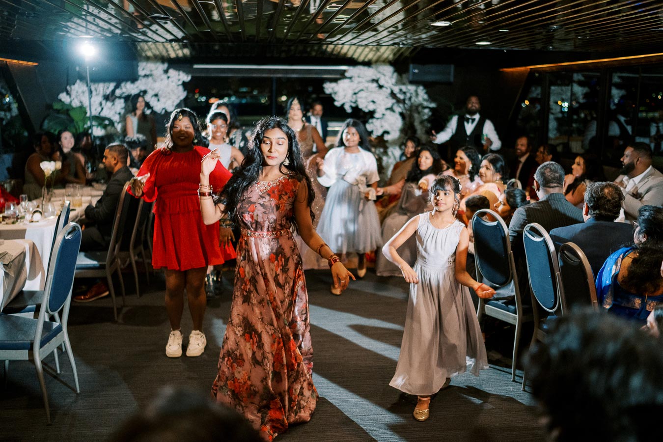 Group of people dancing joyfully at a lively indoor celebration, featuring women and girls in colorful dresses, surrounded by seated guests at elegantly set tables.