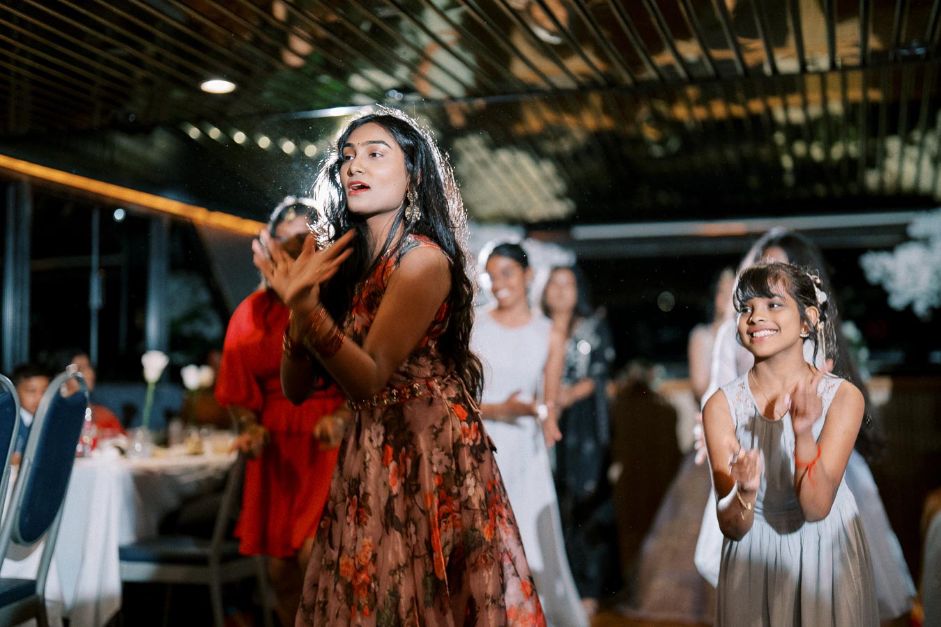 A group of people enjoying a lively dance at an indoor celebration, with a focus on a woman in a floral dress clapping and a smiling young girl in a white dress.