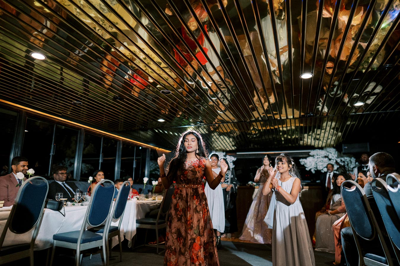 A joyful wedding reception scene with a woman dancing energetically in the center, surrounded by guests seated at tables. The venue has a reflective ceiling, adding a vibrant ambiance. Guests are smiling and engaged, celebrating the occasion.