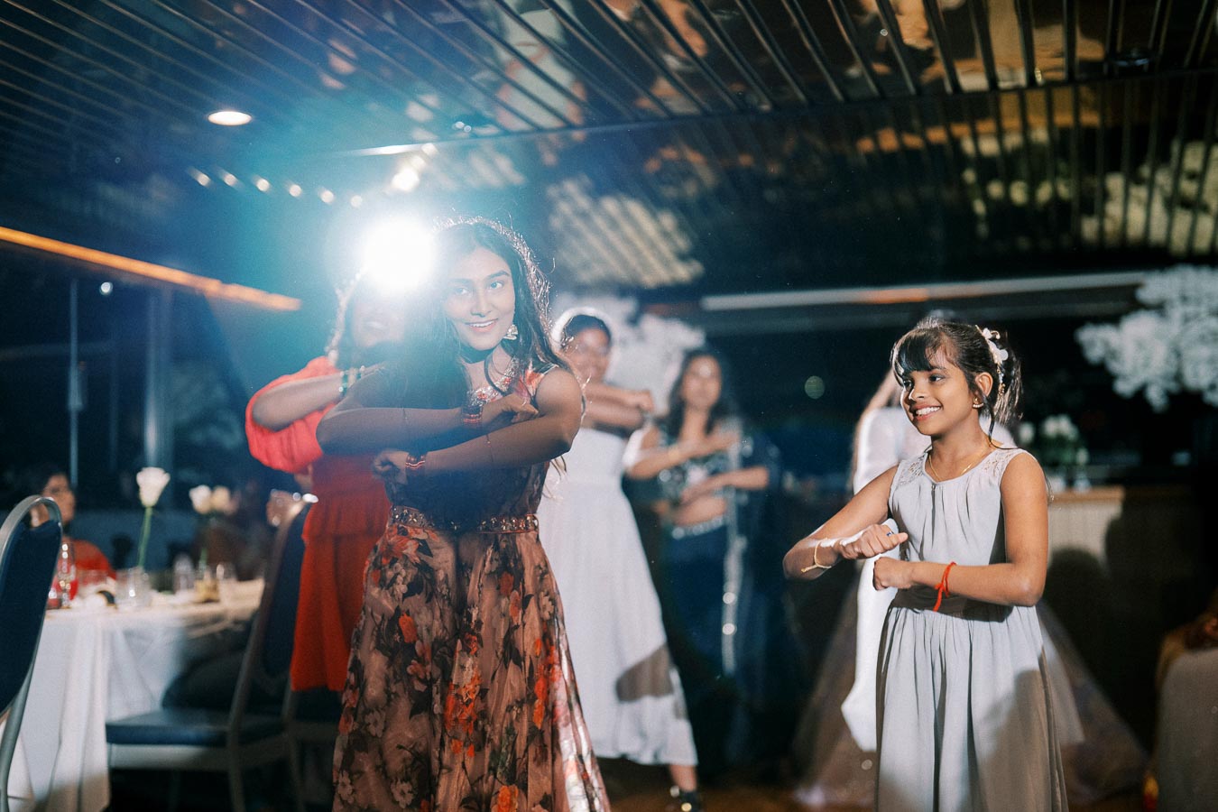 A group of young women and girls dancing joyfully at an indoor celebration, wearing colorful dresses under soft lighting.