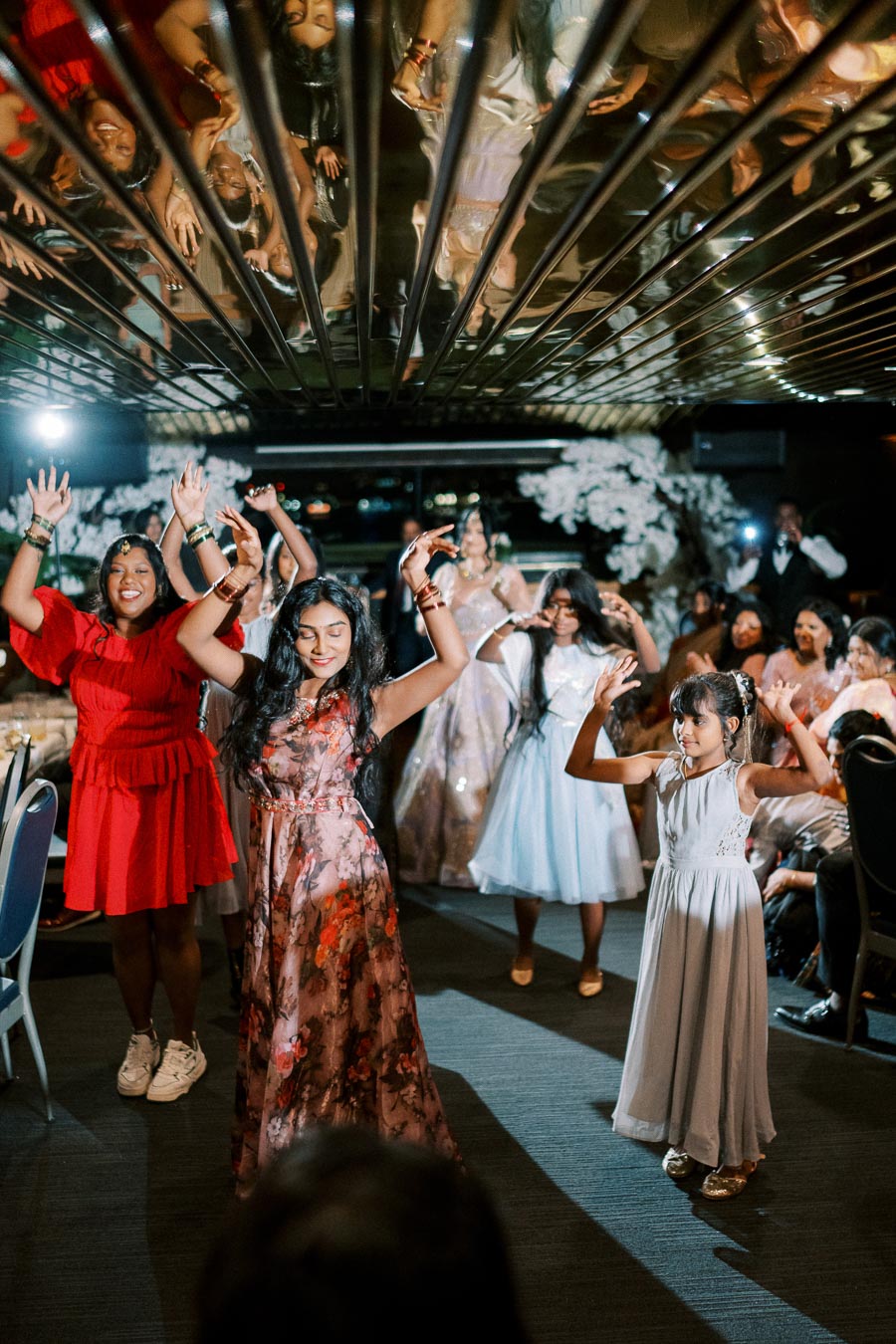 A group of women and girls joyfully dancing at a festive celebration, wearing colorful dresses and accessories, with floral decorations in the background and a mirrored ceiling reflecting the lively scene.