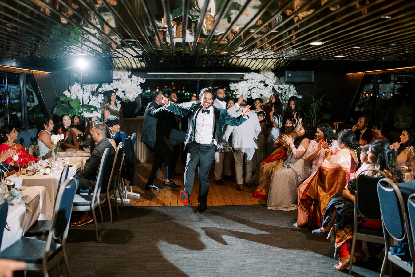 A joyful groom dances in a festive reception hall filled with guests seated at decorated tables, capturing a lively wedding celebration.
