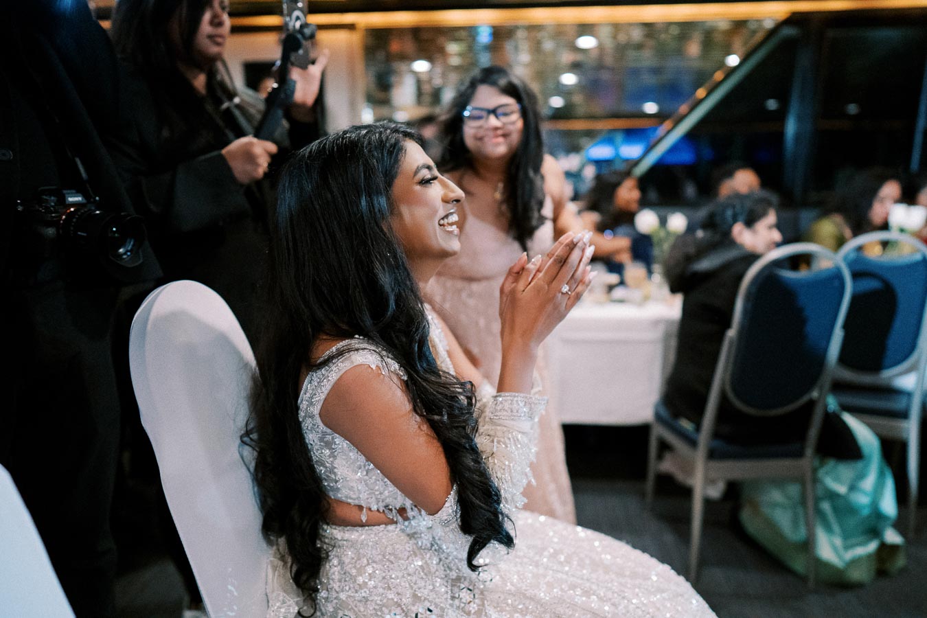 A woman in an elegant white dress with intricate beadwork sits at a table, smiling and clapping her hands. She appears to be enjoying a festive indoor event, surrounded by seated guests, with a photographer and a woman standing nearby.