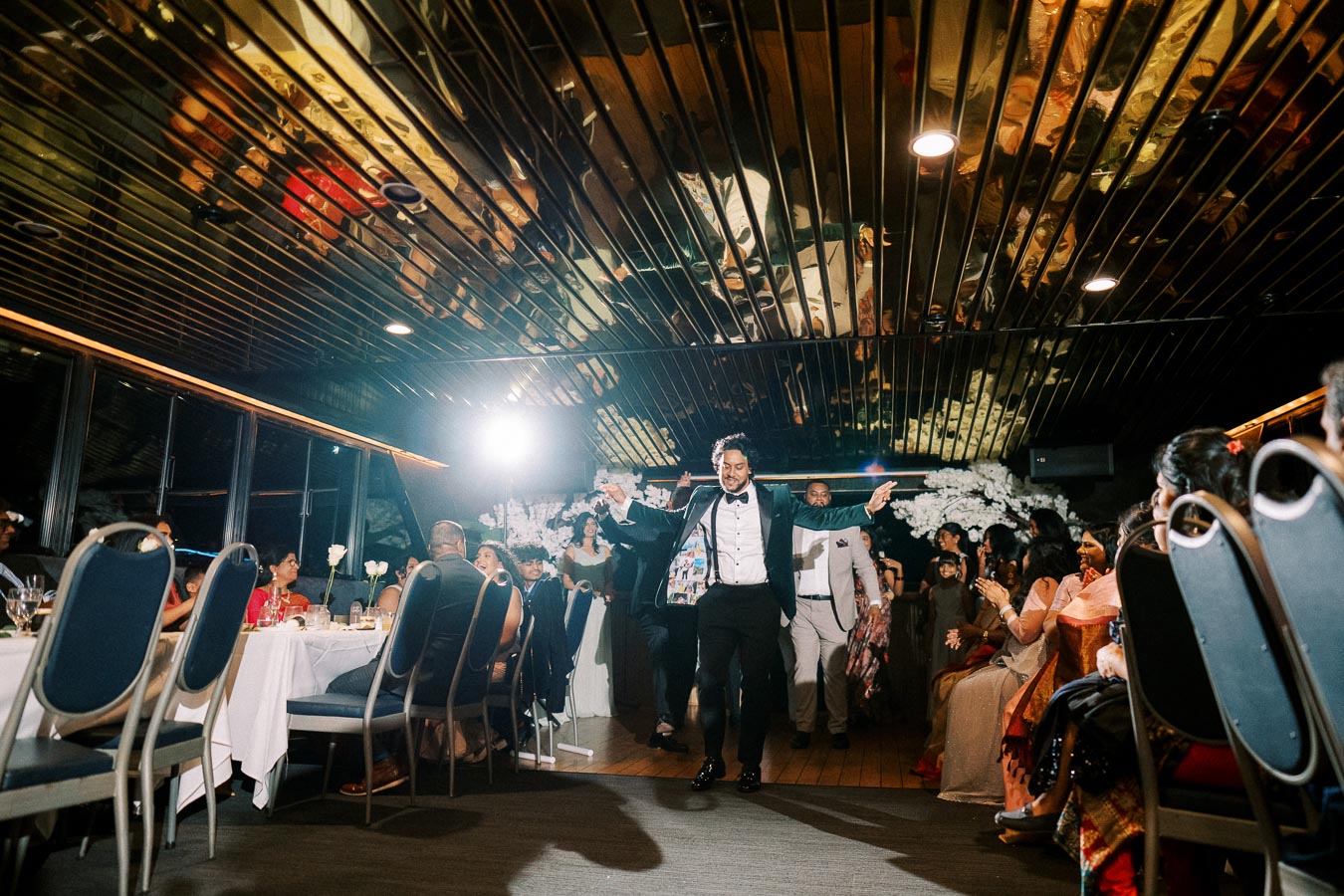 A groom in a tuxedo energetically enters a festive wedding reception, with guests applauding and celebrating in an elegantly decorated venue.