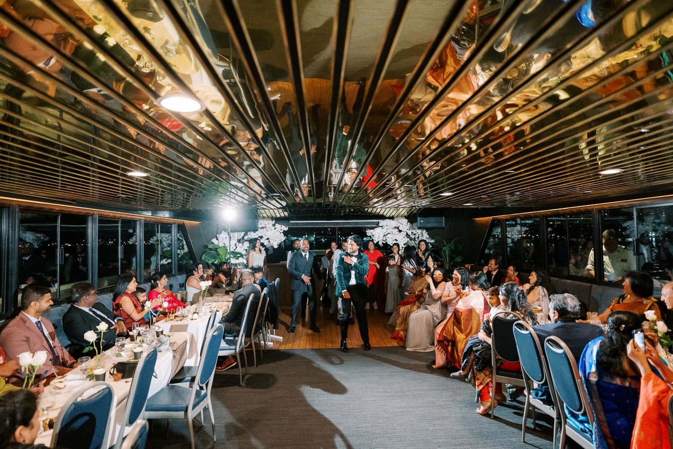 Guests enjoying a lively wedding reception on a beautifully decorated modern boat, with a reflective ceiling and elegant floral arrangements, creating a festive and joyful atmosphere.