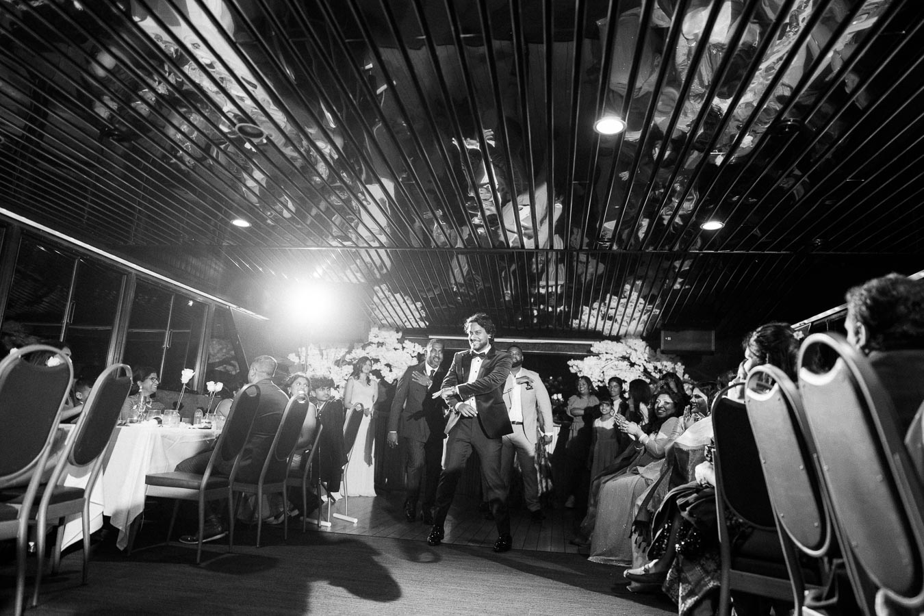 Black and white image of a lively wedding reception, featuring a man in a suit dancing in the center of a modern venue, surrounded by seated guests who are clapping and smiling. The ceiling reflects the scene, enhancing the festive atmosphere.