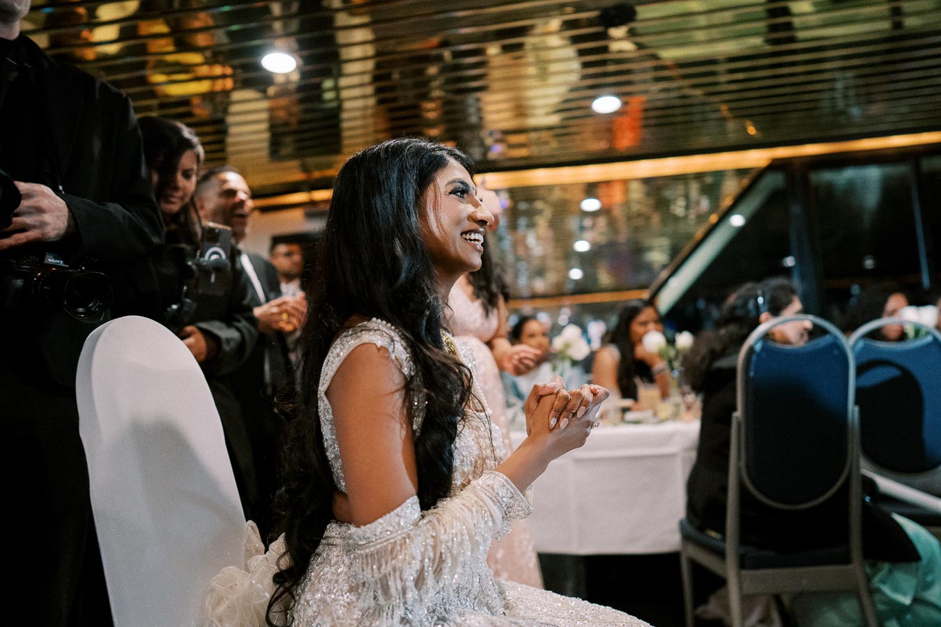 A smiling woman in an elegant, sparkling dress sits delightedly during a formal event, surrounded by people and festive decor.