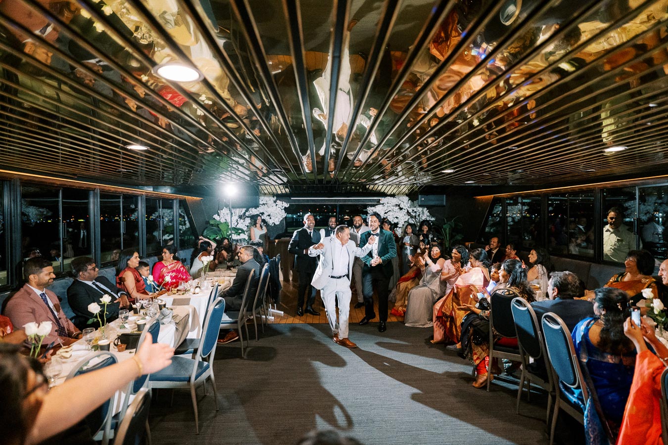 Wedding reception on a boat with guests cheering and clapping as the groom, wearing a light suit, dances down the aisle. The venue features elegant decor with flowers and a reflective ceiling.