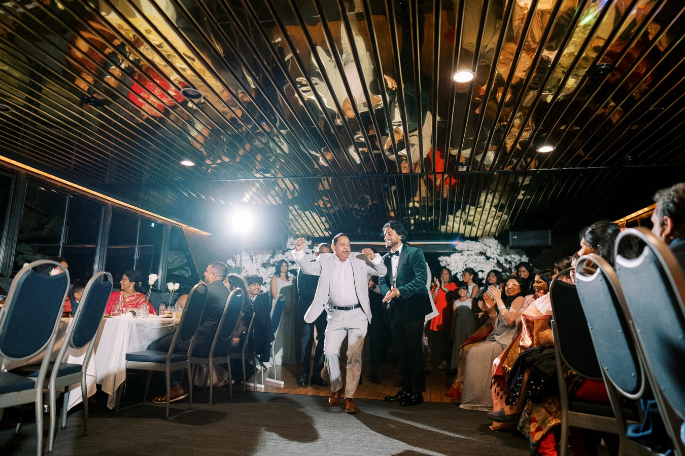 A joyful moment captured during a lively wedding reception, featuring elegantly dressed guests seated at tables, with two men dancing exuberantly in the aisle.
